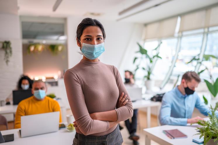 Confident Indian female office worker standing with crossed arms in an office wearing a protective face mask. She is looking at camera.